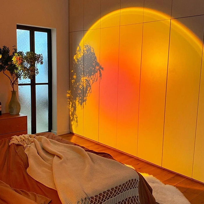 The lamp is used to create a cozy, warm light effect on a bedroom wall, with a gradient transitioning from yellow to orange. The image emphasizes the lamp's capability to create a relaxing ambiance in a home setting.