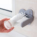 A person cleans a glass by rotating it against a glass cleaner brush that is mounted horizontally on a tiled kitchen wall using suction cups. The brush has stiff white bristles to scrub the inside of the glass.