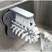 A double-sided glass cleaner brush with stiff white bristles is shown attached to a kitchen sink. The brush is ready to be used for cleaning glasses.