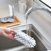 A person rinses a white glass cleaning brush under a running faucet in a kitchen sink. The brush has multiple bristles and is held by the handle. In the background, another brush is seen attached to a wooden board on the countertop.