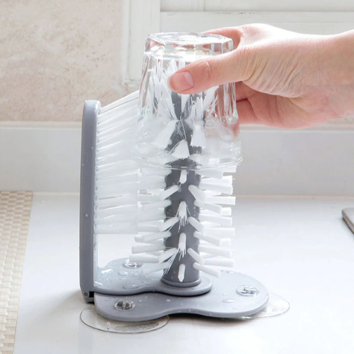 A glass is being cleaned by sliding it over a vertical, bristle-covered glass cleaner brush mounted on the kitchen counter. The brush's bristles thoroughly scrub the inside of the glass.