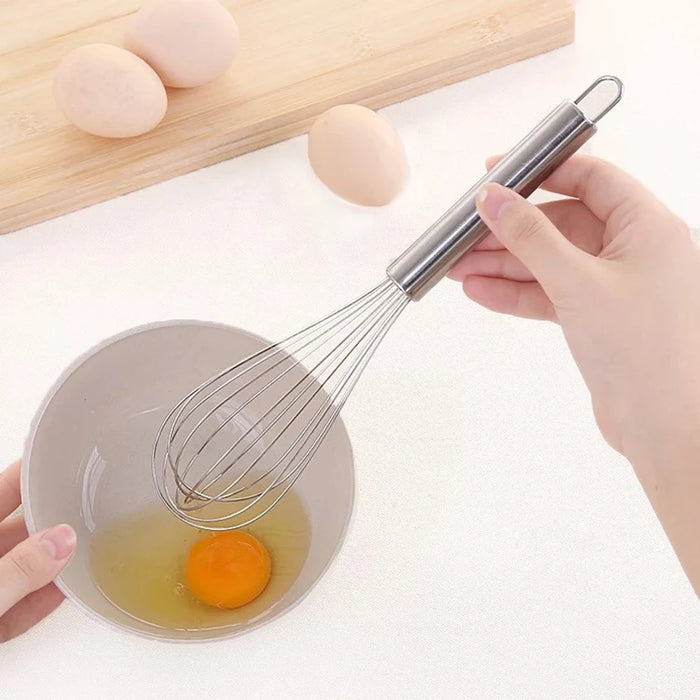 A hand is holding a stainless steel whisk over a small bowl containing a cracked egg. There are more eggs in the background on a wooden cutting board, indicating the whisk is being used for egg preparation.