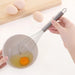 A hand is holding a stainless steel whisk over a small bowl containing a cracked egg. There are more eggs in the background on a wooden cutting board, indicating the whisk is being used for egg preparation.