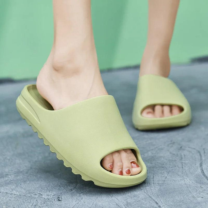 A close-up of a person's feet wearing light green slide sandals on a concrete surface, with a green background.