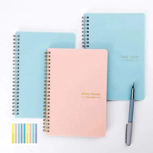 Three spiral-bound weekly planners with pastel-colored covers (two light blue and one pink) and a pen placed next to them. There is also a small color palette at the bottom left corner showing various colors.