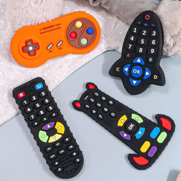 Four silicone teethers shaped like remote controls and a game controller in various designs and colors laid out on a soft surface.