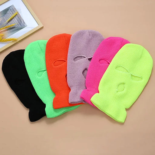 A selection of knitted ski masks in various bright colors, laid flat side by side. The colors include black, neon green, orange, lavender, hot pink, and neon yellow. Each mask features two eye holes and one mouth hole.