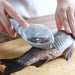 Another image of a person scaling a fish with the tool, with a focus on the built-in cover collecting the scales.