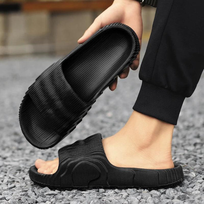 A close-up of a person wearing black slide sandals, holding one sandal in hand with the other foot in the other sandal on a gravel surface.