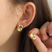 a woman wearing gold stud earrings with an abstract, irregular shape. The earrings have a smooth, reflective surface, adding a modern and artistic element to her style. She holds one earring near her ear to show the detail.