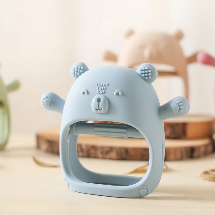 A close-up of a blue silicone teether shaped like a bear, placed on a wooden surface with blurred toys in the background.