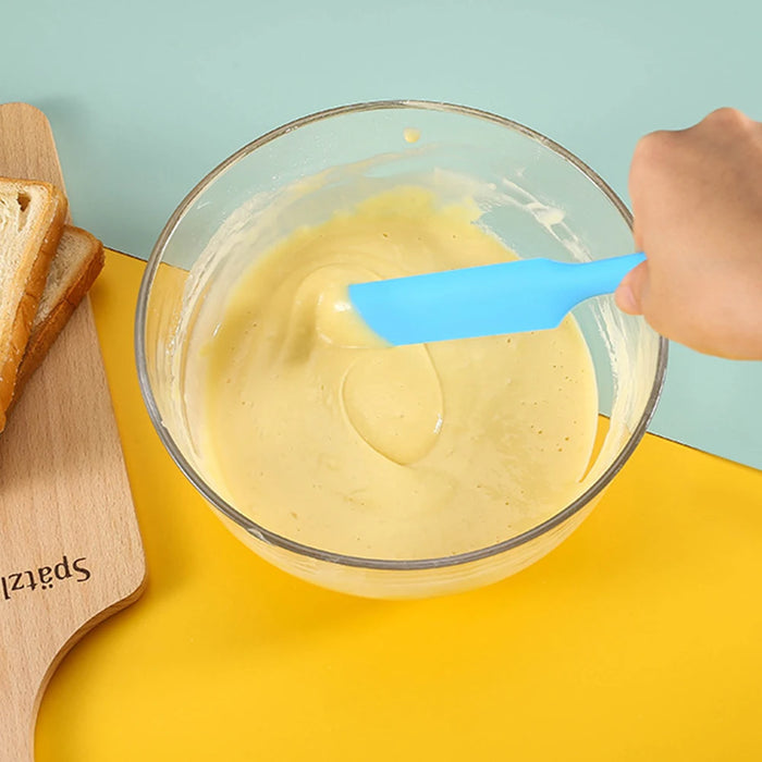 a person using a bright blue spatula to mix a creamy batter in a clear glass bowl, placed on a colorful background with slices of bread nearby.