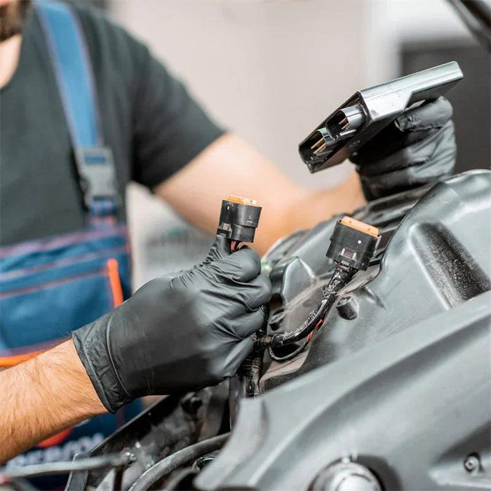 A close-up image shows a mechanic working on a vehicle. The mechanic is wearing black gloves and handling electrical connectors, demonstrating the protective nature of the gloves in a hands-on work environment.