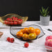 A variety of wire fruit baskets in black, white, and gold, each containing different fruits like oranges, tomatoes, and apples. The arrangement is on a marble surface, emphasizing a fresh and stylish kitchen or dining room display.