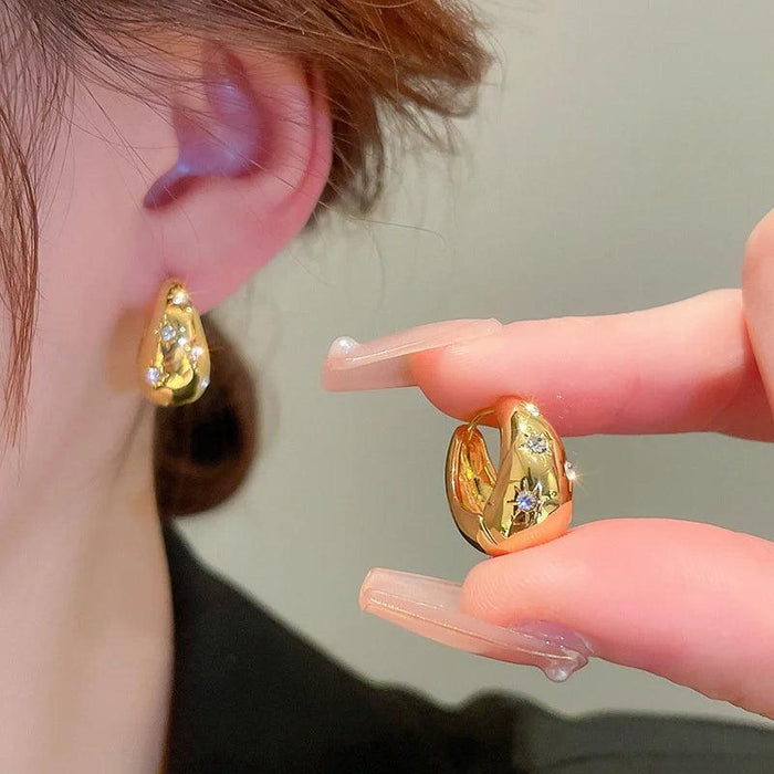 a woman wearing gold earrings with embedded crystals. The design is small and chunky, with a sparkling detail that adds a touch of glamour. She holds one earring close to her ear to show the intricate design.