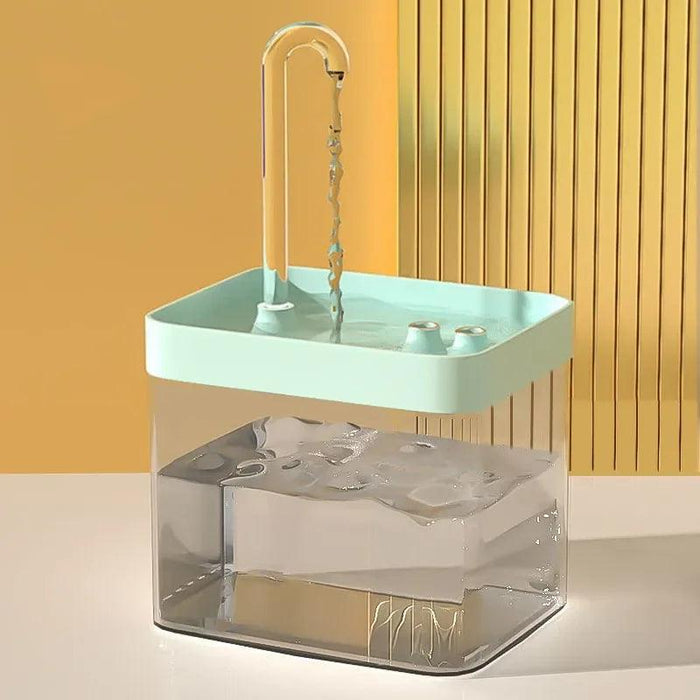 A pet water fountain with a turquoise top and a clear base, filled with water and a clear curved spout extending from the top. It is set against a beige background.