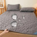 Gray quilted mattress cover pad on a bed, showing water droplets on the surface to highlight the waterproof feature. Matching pillows are on the bed, with a cozy room setup including a small table and a plant.