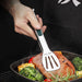 Stainless steel tongs being used to flip a piece of meat in a frying pan with vegetables.