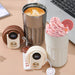 Two thermal coffee mugs, one stainless steel and one white, placed next to a laptop. The stainless steel mug contains a latte, while the white mug holds a dessert topped with pink whipped cream. Two detachable lids with digital temperature displays are shown in the foreground.