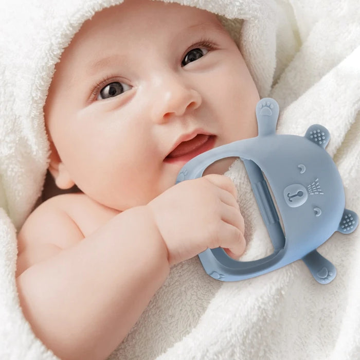 A baby wrapped in a towel holding and chewing on a blue silicone teether shaped like a bear.