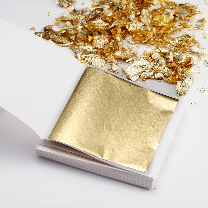 Single gold leaf sheet in a booklet, surrounded by scattered gold flakes for crafting use.