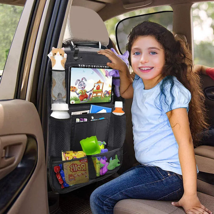 Girl in a car with a black backseat organizer holding a tablet, toys, snacks, and bottles.