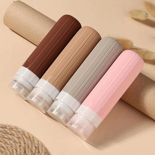 Set of four colorful, silicone travel bottles arranged diagonally on a beige background. The bottles come in shades of brown, beige, gray, and pink, each capped with a clear, twist-off top, perfect for storing liquids securely during travel.