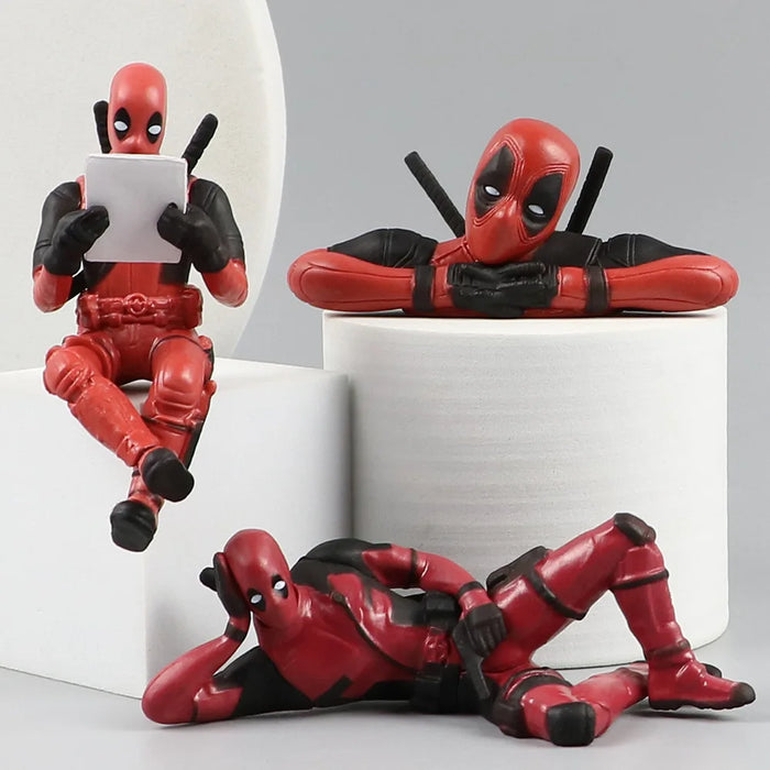 three Deadpool figurines. One is sitting and reading a blank piece of paper, another is resting its chin on its crossed arms, and the third is lying on its side in a relaxed pose, all placed on round white platforms.