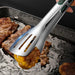 Stainless steel tongs holding a piece of corn on the cob over a grill with other food items cooking.