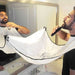 A man trimming his beard over a sink, with a white beard trimming bib attached to the mirror.