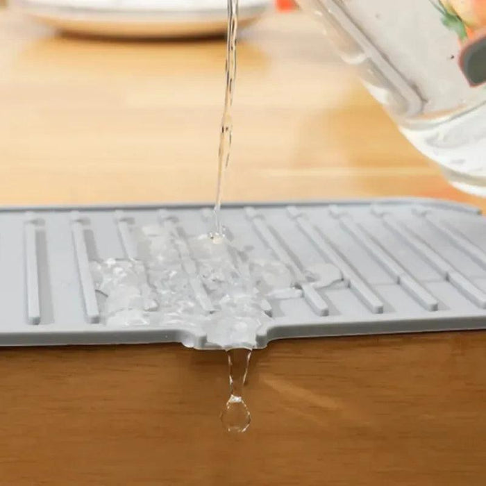 water being poured onto a gray silicone splash guard. The water is shown flowing down the mat, illustrating its function in guiding excess water off surfaces and back into the sink.