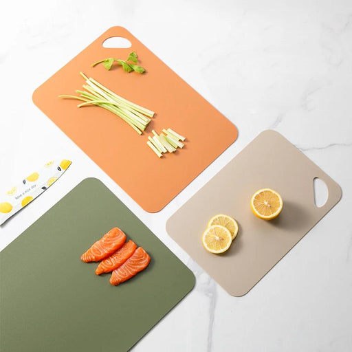 An assortment of colorful cutting boards arranged on a marble countertop. From left to right: an olive green board with sliced salmon, an orange board with fresh celery sticks and basil, and a taupe board with sliced lemons. Each board features a practical handle for easy lifting and storage.