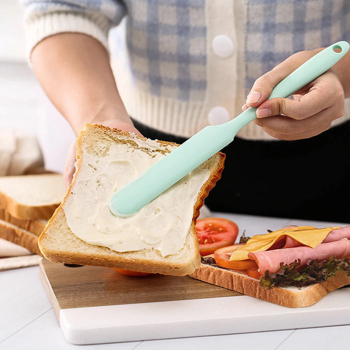 a person spreading butter or cream cheese on a slice of bread using a mint green spatula, preparing a sandwich with various ingredients in the background.