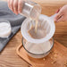 A person is pouring a glass of liquid through a white strainer into a glass bowl, with another glass of liquid on a wooden table.