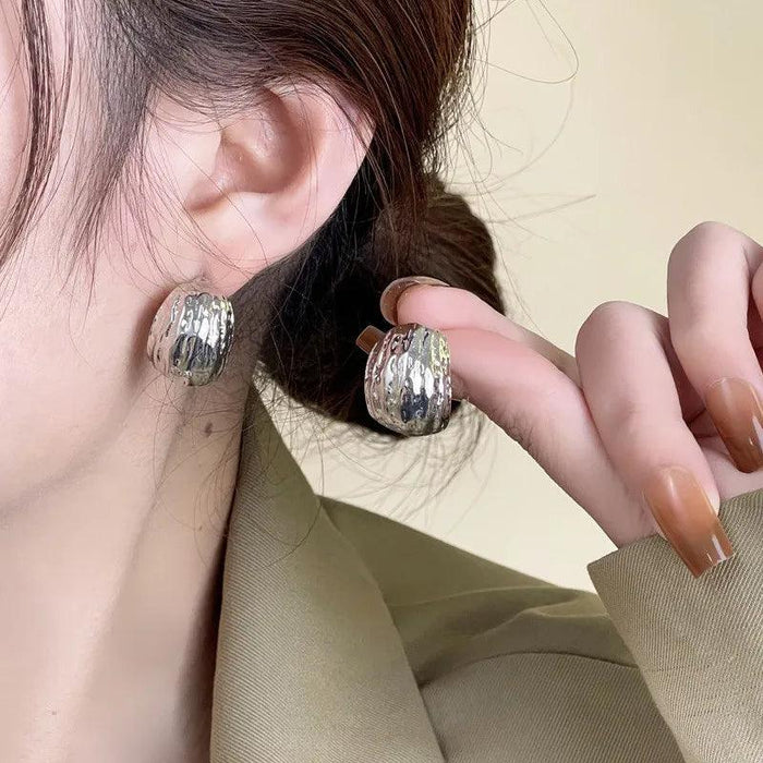 a woman wearing textured silver hoop earrings. The earrings have a hammered finish, giving them a unique and artisanal look. She holds one earring close to her ear to highlight the texture.