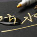 A close-up of a ceramic tile marker writing gold ink on black paper, demonstrating the marker's water-resistant properties.