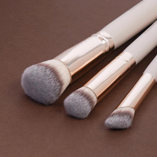 Three makeup brushes with white handles and rose gold ferrules, laid flat on a brown background, showcasing the varying sizes and shapes of the brush heads.