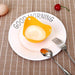 A yellow silicone egg poacher holding a poached egg is placed on a white plate with "GOOD MORNING" written on it. The plate is set on a brown checkered tablecloth, with a fork and spoon beside it.