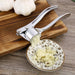 A metal garlic press crushing garlic into a small bowl, with garlic bulbs in the background.