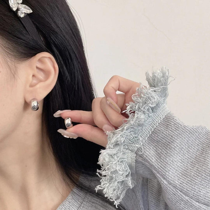 a woman with short dark hair wearing small, chunky silver hoop earrings. The earrings are simple yet chic, complementing her casual outfit. She holds one earring near her ear to showcase the design.