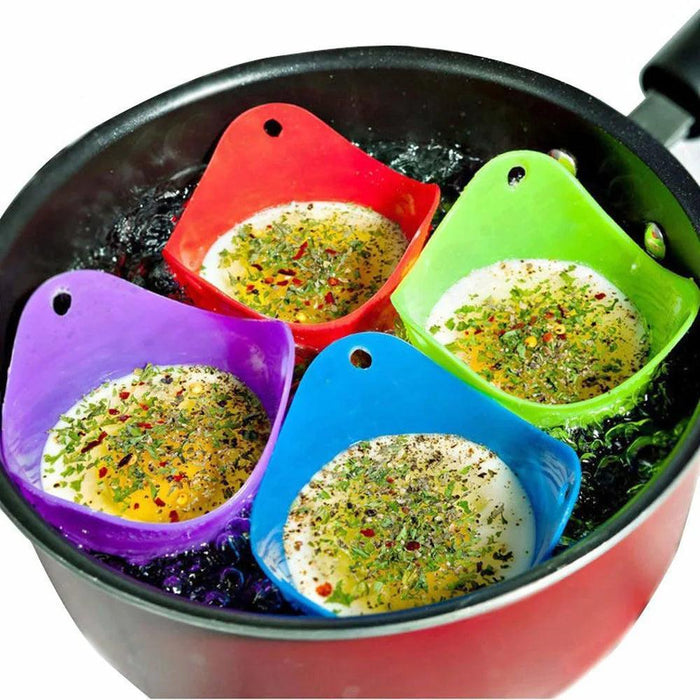 Four silicone egg poachers in red, green, blue, and purple colors, each holding a poached egg topped with herbs and spices, are floating in boiling water inside a black pan.