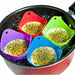 Four silicone egg poachers in red, green, blue, and purple colors, each holding a poached egg topped with herbs and spices, are floating in boiling water inside a black pan.