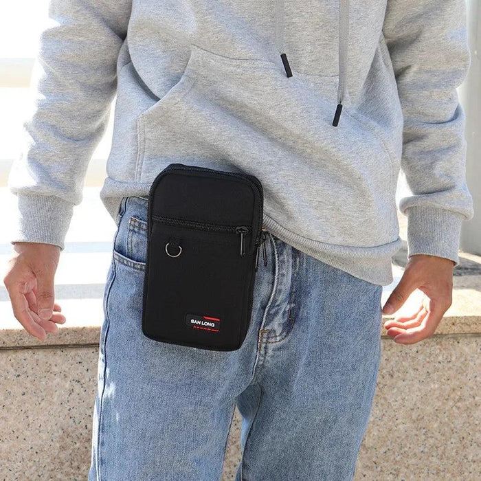 A black crossbody pouch with a front zipper pocket and a small D-ring attachment, worn by a person in blue jeans and a gray hoodie. The pouch is clipped to the waistband of the jeans, demonstrating its portability.