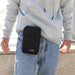 A black crossbody pouch with a front zipper pocket and a small D-ring attachment, worn by a person in blue jeans and a gray hoodie. The pouch is clipped to the waistband of the jeans, demonstrating its portability.