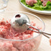 Close-up of stainless steel meatball tongs shaping a meatball from a bowl of ground meat.