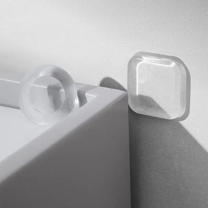 A clear round and a clear square silicone bumper pad are attached to a corner of a white piece of furniture.