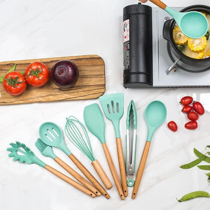 A set of turquoise silicone kitchen utensils placed next to each other on a white marble with tomatoes, cherry tomatoes, purple onions, and green beans in the background. In addition, there is a pot on the stove that cooks corn inside.