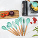 A set of turquoise silicone kitchen utensils placed next to each other on a white marble with tomatoes, cherry tomatoes, purple onions, and green beans in the background. In addition, there is a pot on the stove that cooks corn inside.