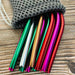 A set of colorful, reusable metal straws, partially in a grey mesh pouch, displayed on a wooden surface.