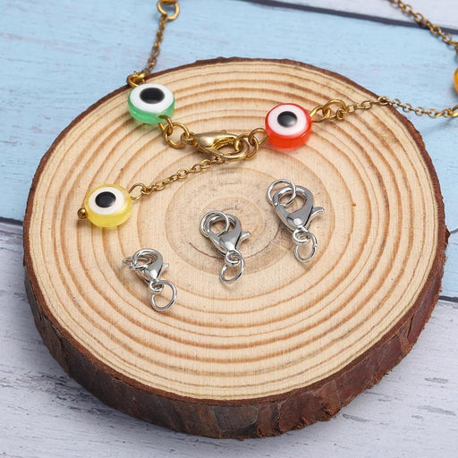 A gold chain bracelet with colorful eye-shaped beads is laid out on a wooden slice, alongside three silver lobster clasps, showcasing their size and detail.